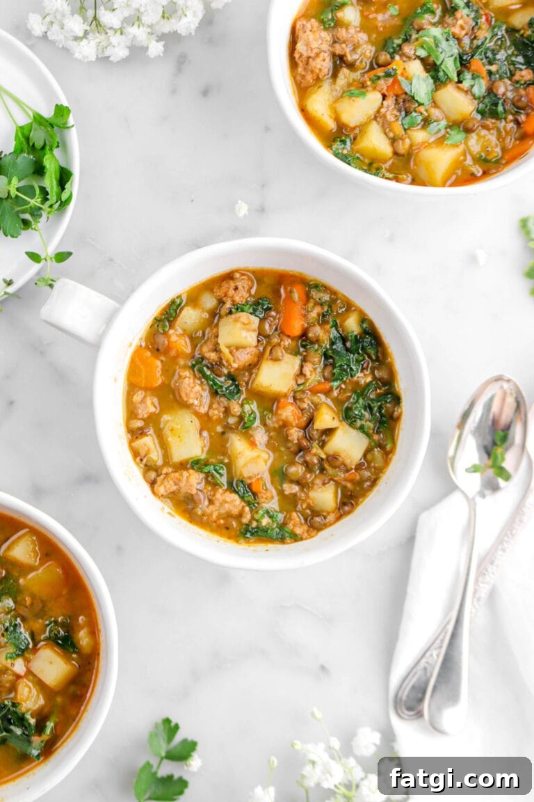 close up of vegan sausage and kale soup with spoons beside, two more bowls of soup, and fresh herbs on marble surface.