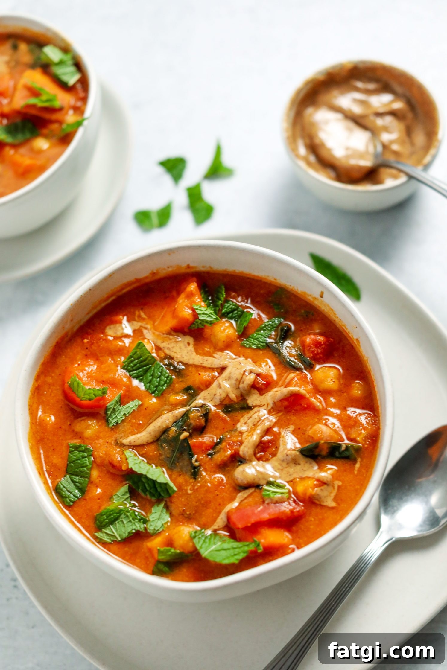 Authentic West African Peanut Stew Recipe 6 African Peanut Butter Stew served in a bowl on a plate with a spoon and fresh mint on top and peanut butter bowl on the side by Flora & Vino