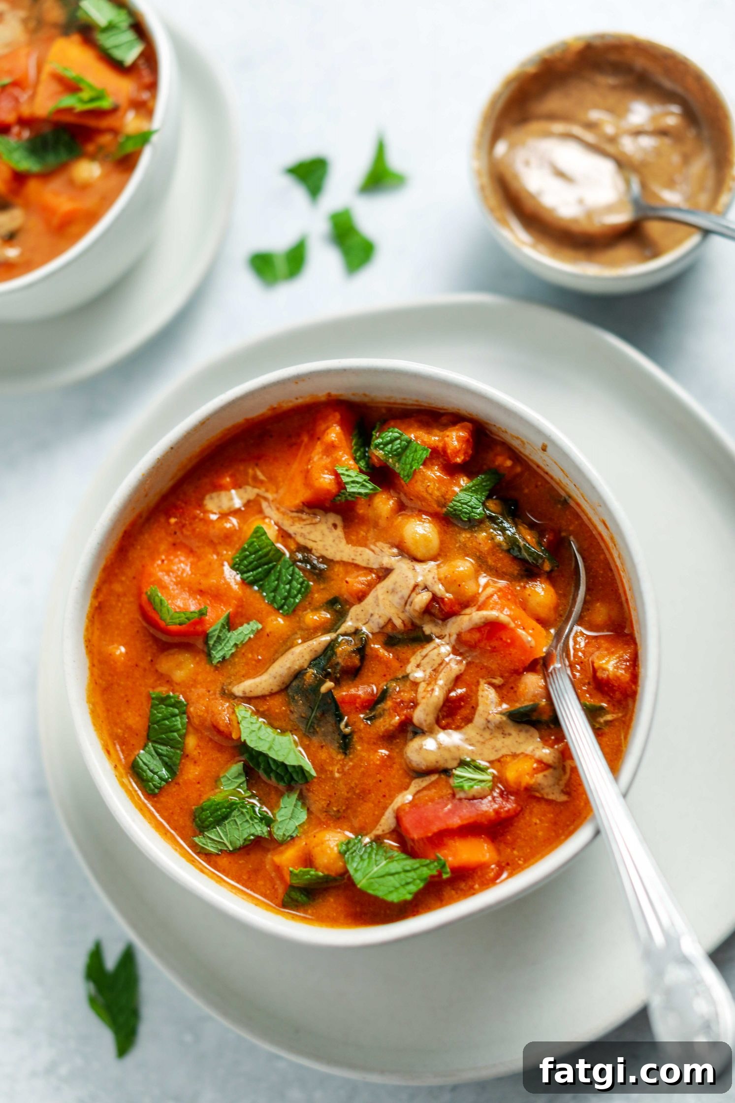 Authentic West African Peanut Stew Recipe 8 African Peanut Butter Stew served in a bowl on a plate with a spoon and fresh mint on top and a bowl of peanut butter on the side by Flora & Vino