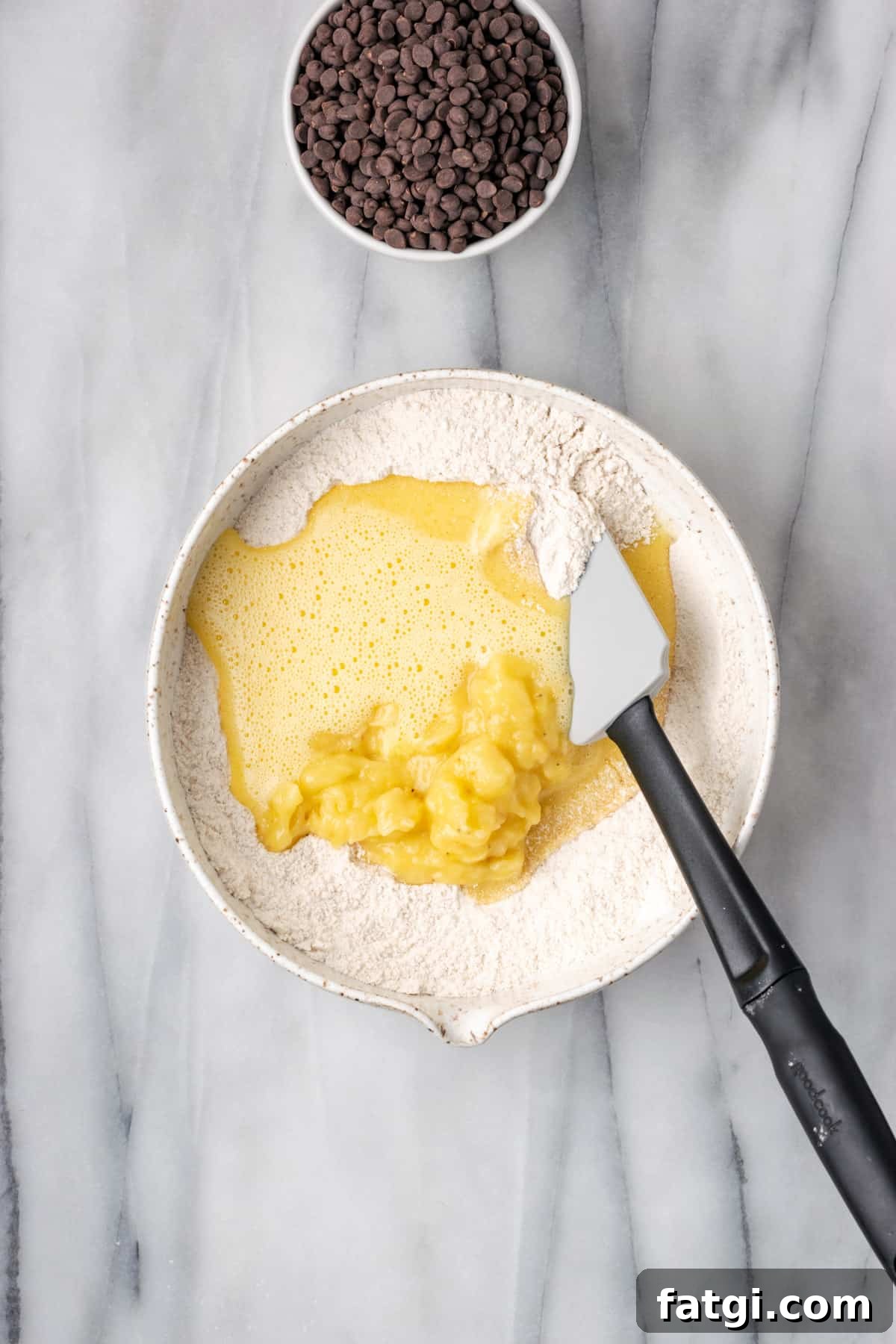 Egg mixture and mashed banana being folded into the dry ingredients in a large white bowl.