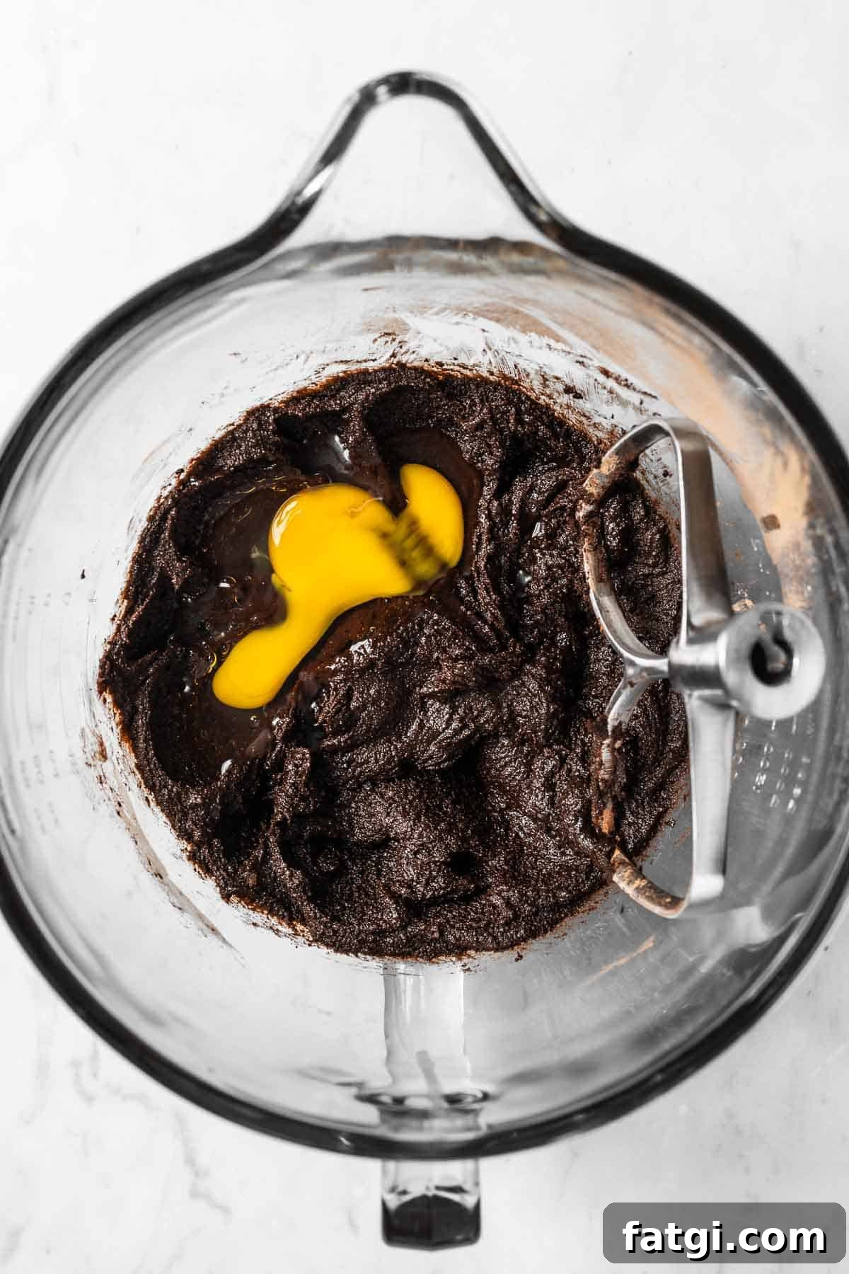 An egg added to melted butter, sugar and cocoa powder in a glass mixing bowl.