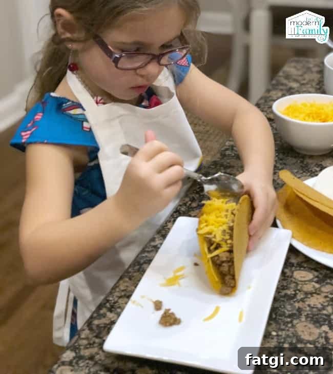 A little girl wearing a white apron sitting at a table putting cheese on a taco.