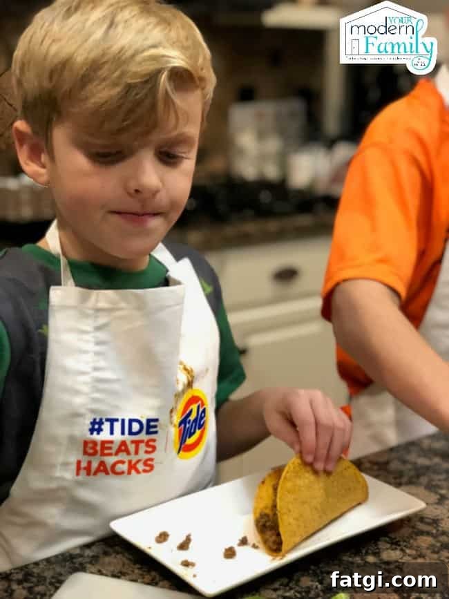 A young boy wearing a white apron sitting at a table putting meat into a taco shell.