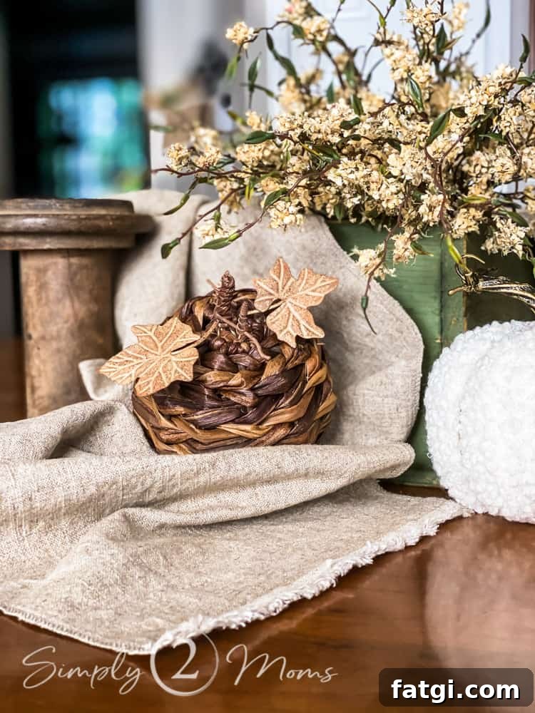 Table with a neutral fall centerpiece made of green wooden vintage caddy filled with faux dried white flowers, a natural linen table runner is tucked inside and trails over the edges, In front, there is a vintage wooden spool, a cream sweater pumpkin, a woven pumpkin, and a white fleece pumpkin.