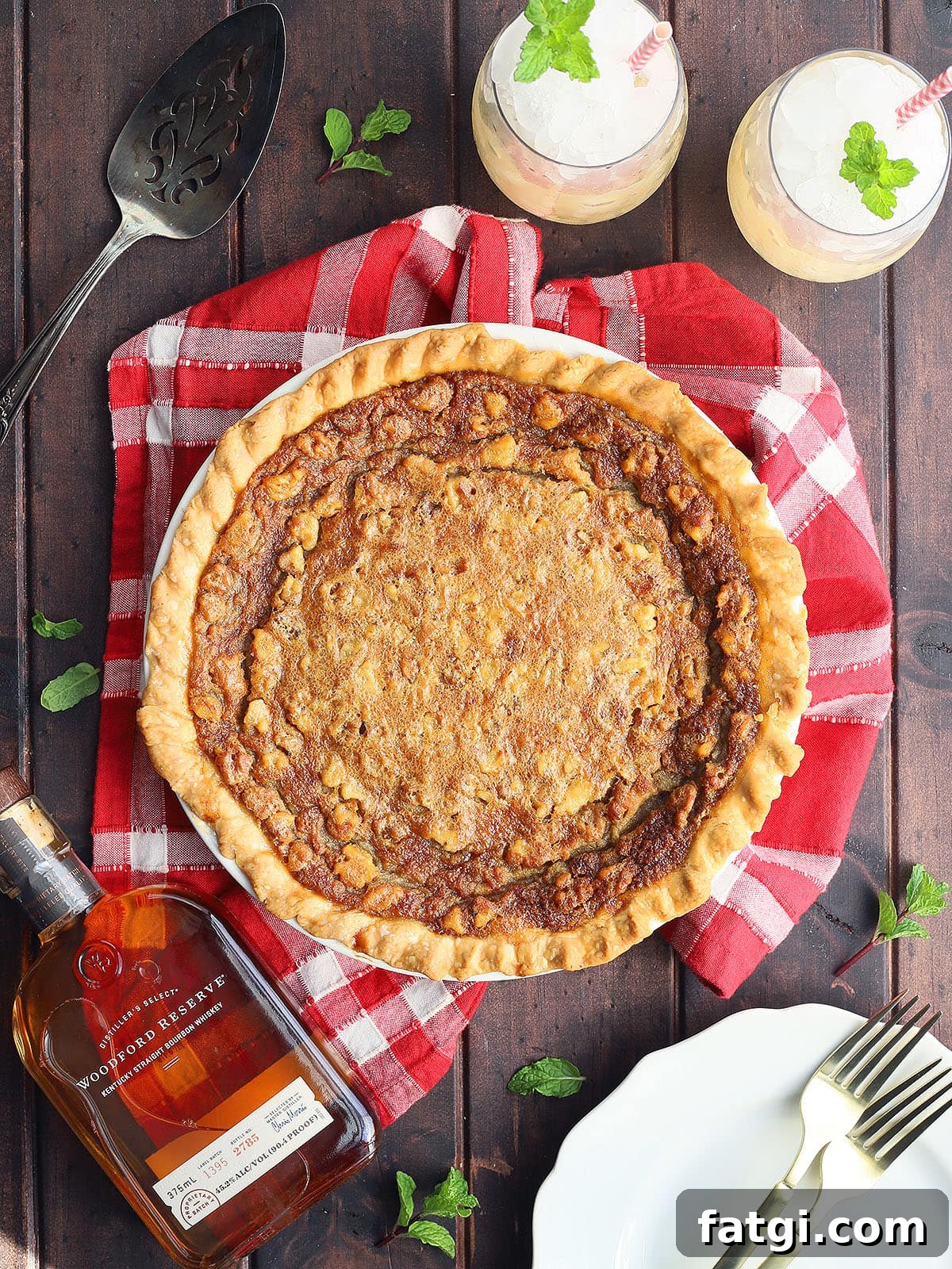 Whole Kentucky Derby pie on a dark wooden background with plates, forks, two mint juleps and a bottle of bourbon to the side.