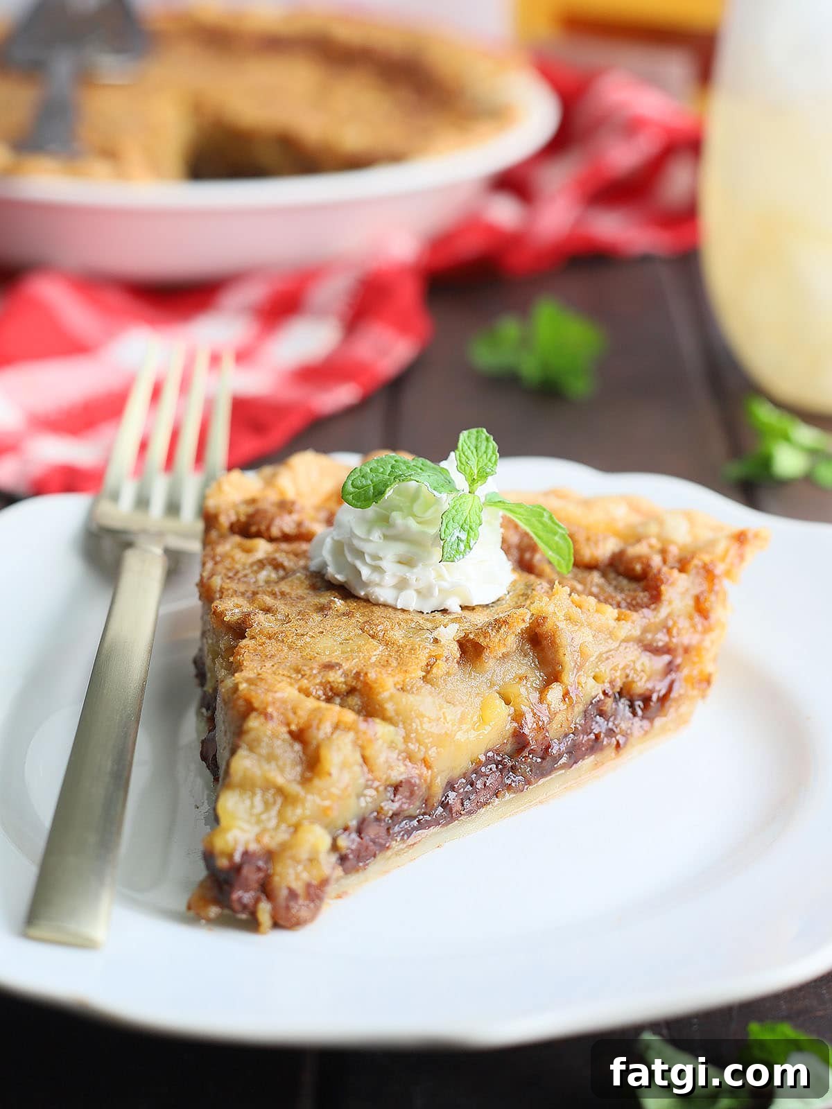 One slice of Kentucky Derby pie garnished with whipped cream and a mint sprig on a white plate with a gold fork to the side and pie in the background.