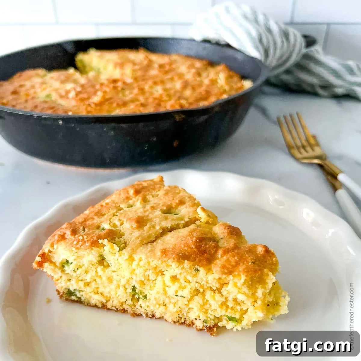 Jiffy Mix Jalapeno Cheddar Cornbread Recipe 5 a white plate with a slice of jalapeno cornbread with a skillet with cornbread, bread knife, fork, and a striped cloth in the background.