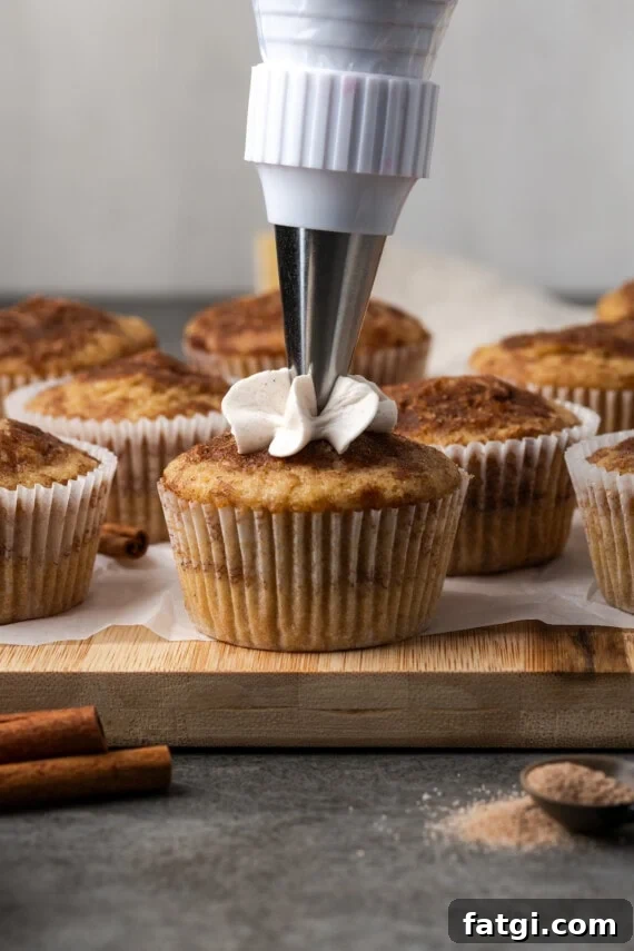 A piping tip pipes cinnamon frosting onto a snickerdoodle cupcake, with more unfrosted cupcakes in the background.