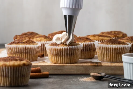 A piping tip pipes cinnamon frosting into the beginning of a swirl on a snickerdoodle cupcake, with more unfrosted cupcakes in the background.