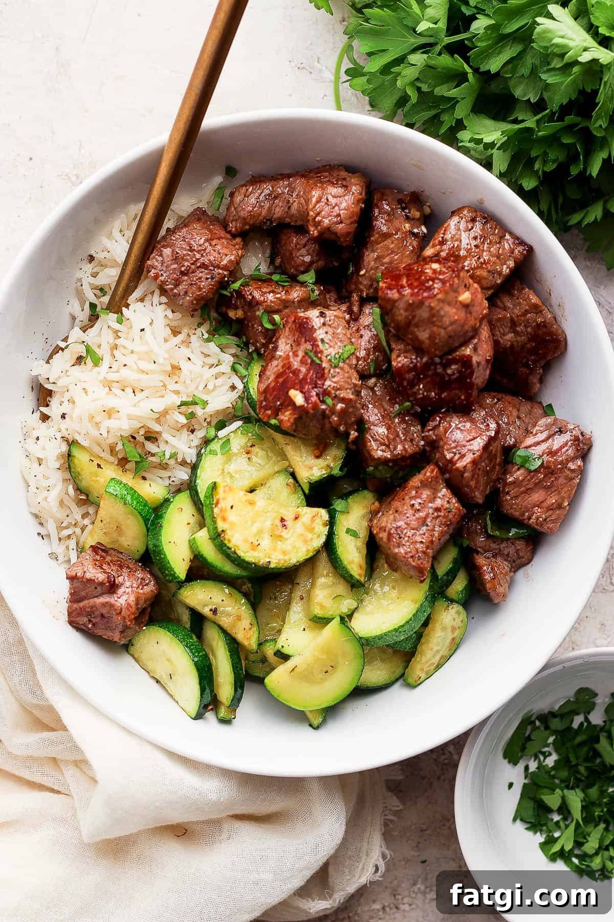 Garlic Butter Steak Bites Recipe 8 Garlic butter steak bites in a white bowl with white rice and zucchini.