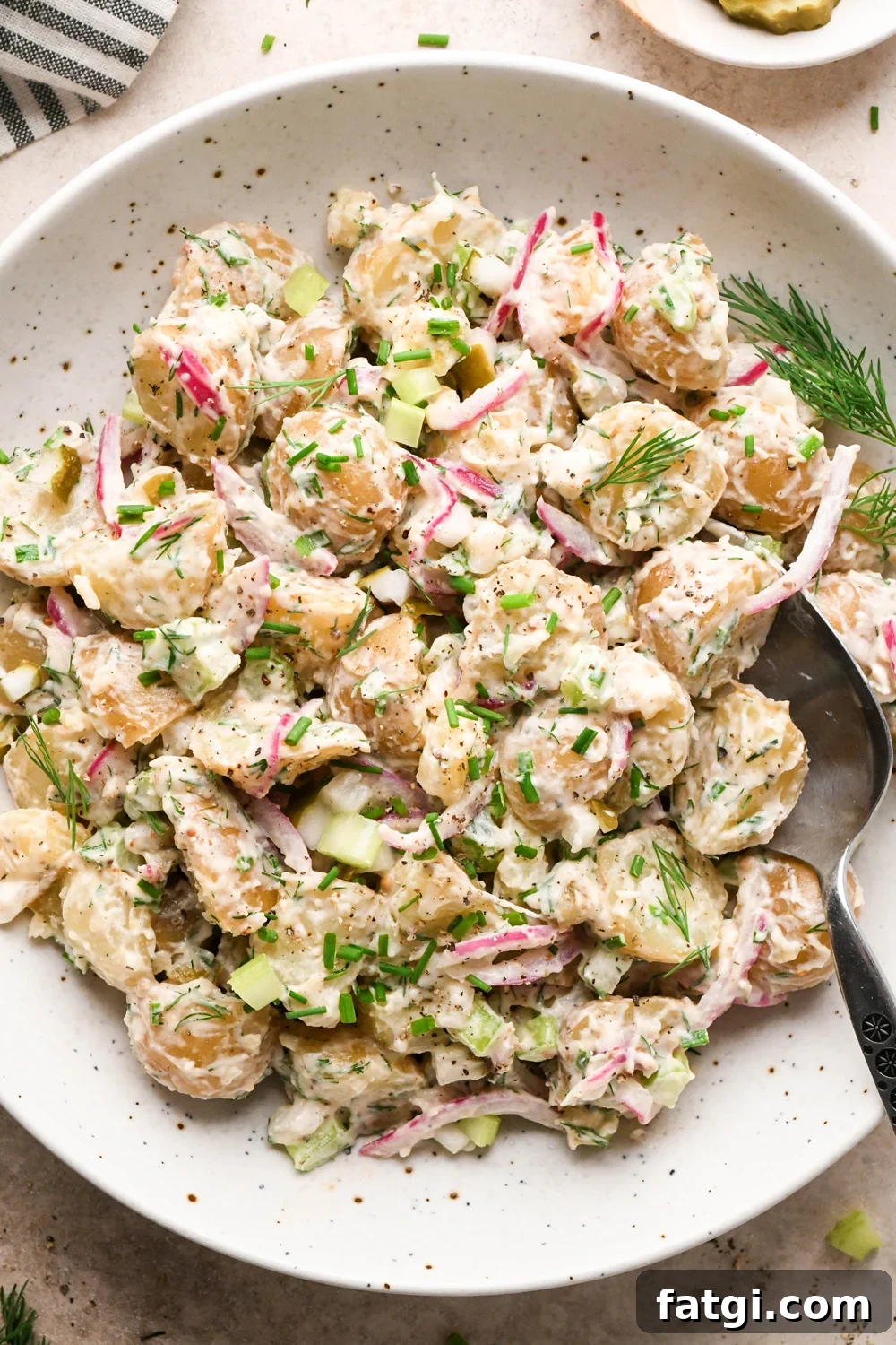 A large shallow speckled bowl filled with creamy mayo-based dill pickle potato salad. A spoon is angled into the bowl, and the potato salad is garnished with fresh dill, fresh snipped chives, and black pepper.