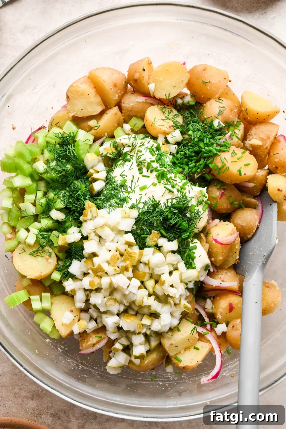 Mayonnaise, chopped pickles, diced celery, and fresh herbs added to the large glass mixing bowl with potatoes, before stirring to combine.