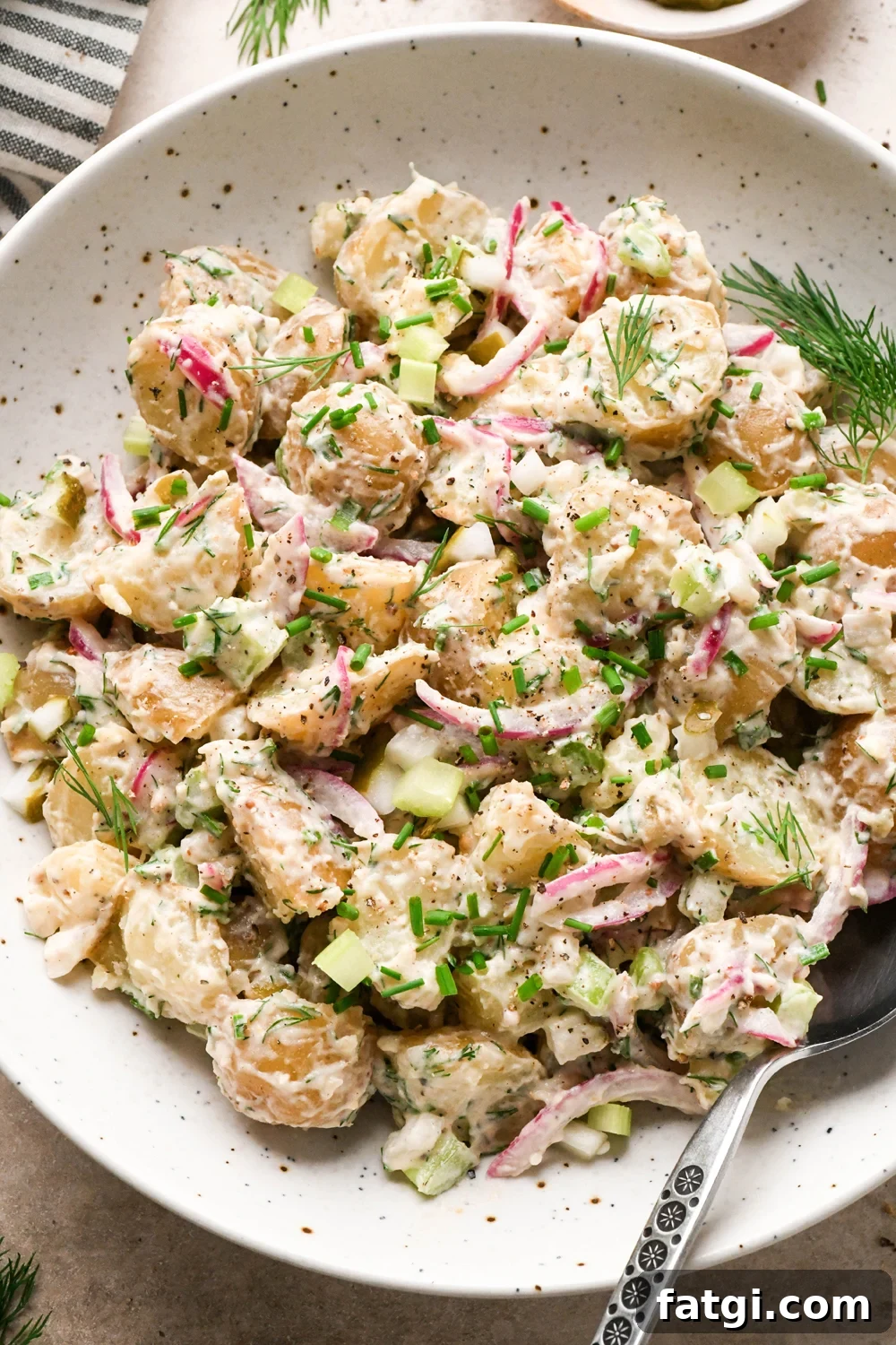 A large shallow speckled bowl filled with creamy mayo-based dill pickle potato salad. A spoon is angled into the bowl, and the potato salad is garnished with fresh dill, fresh snipped chives, and black pepper.