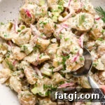 A large shallow speckled bowl filled with creamy mayo-based dill pickle potato salad with a spoon lifting out a portion to show the texture. The potato salad is garnished with fresh dill, fresh snipped chives, and black pepper.