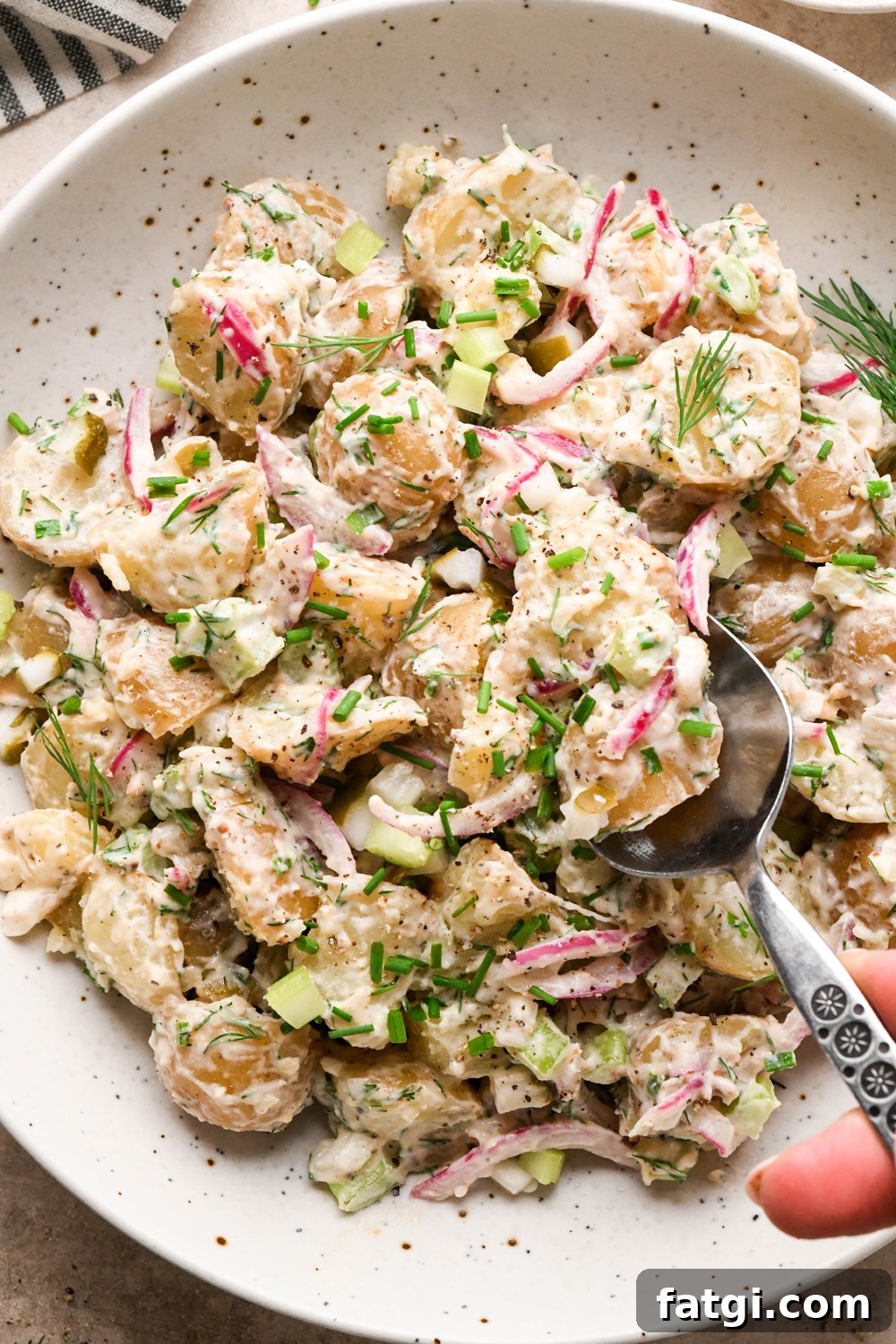 A large shallow speckled bowl filled with creamy mayo-based dill pickle potato salad with a spoon lifting out a portion to show the texture. The potato salad is garnished with fresh dill, fresh snipped chives, and black pepper.