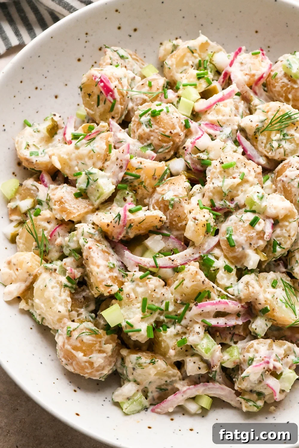 A large shallow speckled bowl filled with creamy mayo-based dill pickle potato salad. The potato salad is garnished with fresh dill, fresh snipped chives, and black pepper.