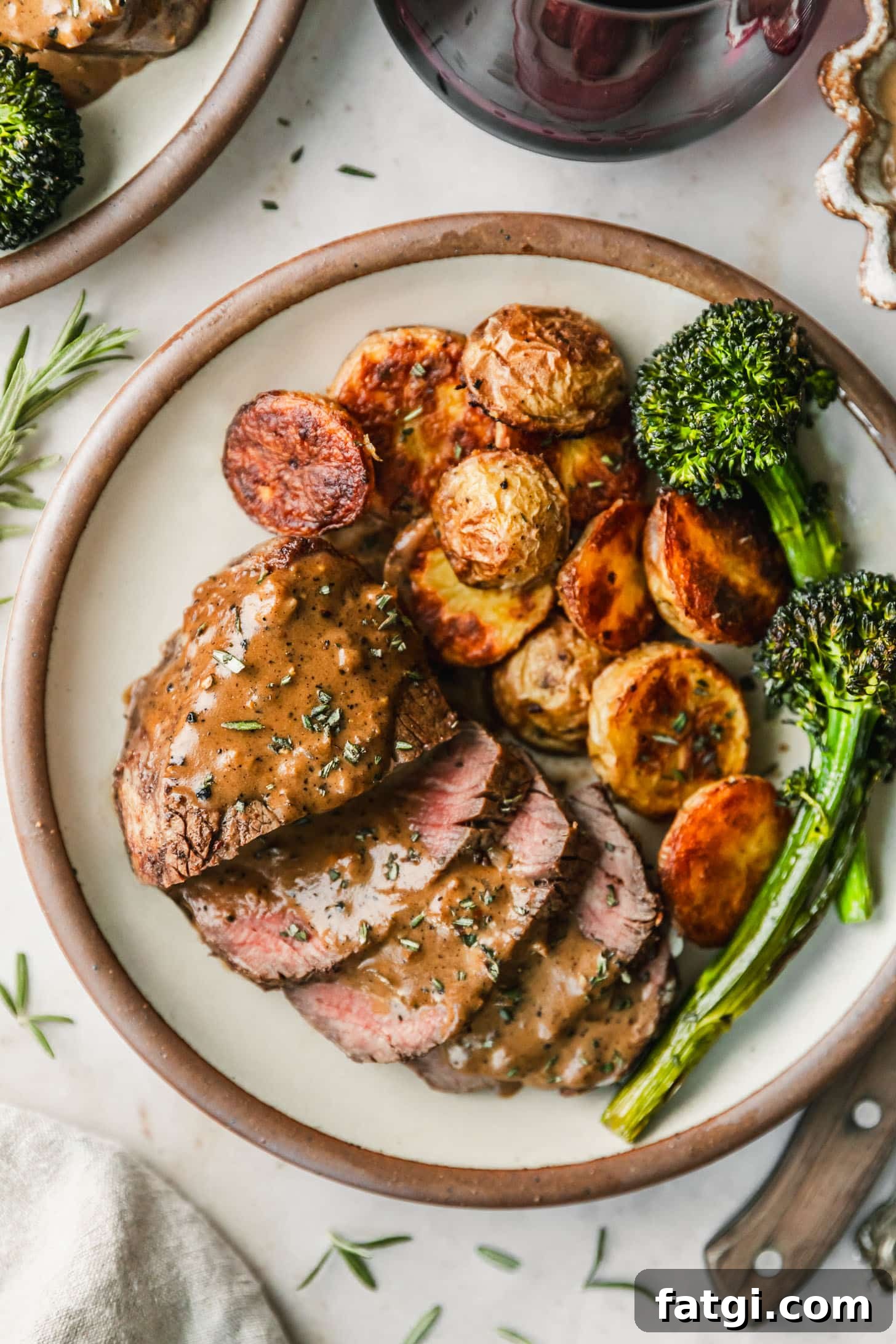 Reverse-Seared Filet Mignon with Creamy Peppercorn Sauce 2 A white plate with potatoes, broccolini, and reverse sear filet mignon with peppercorn sauce on a beige marble counter next to a wood knife, beige linen, and red wine.