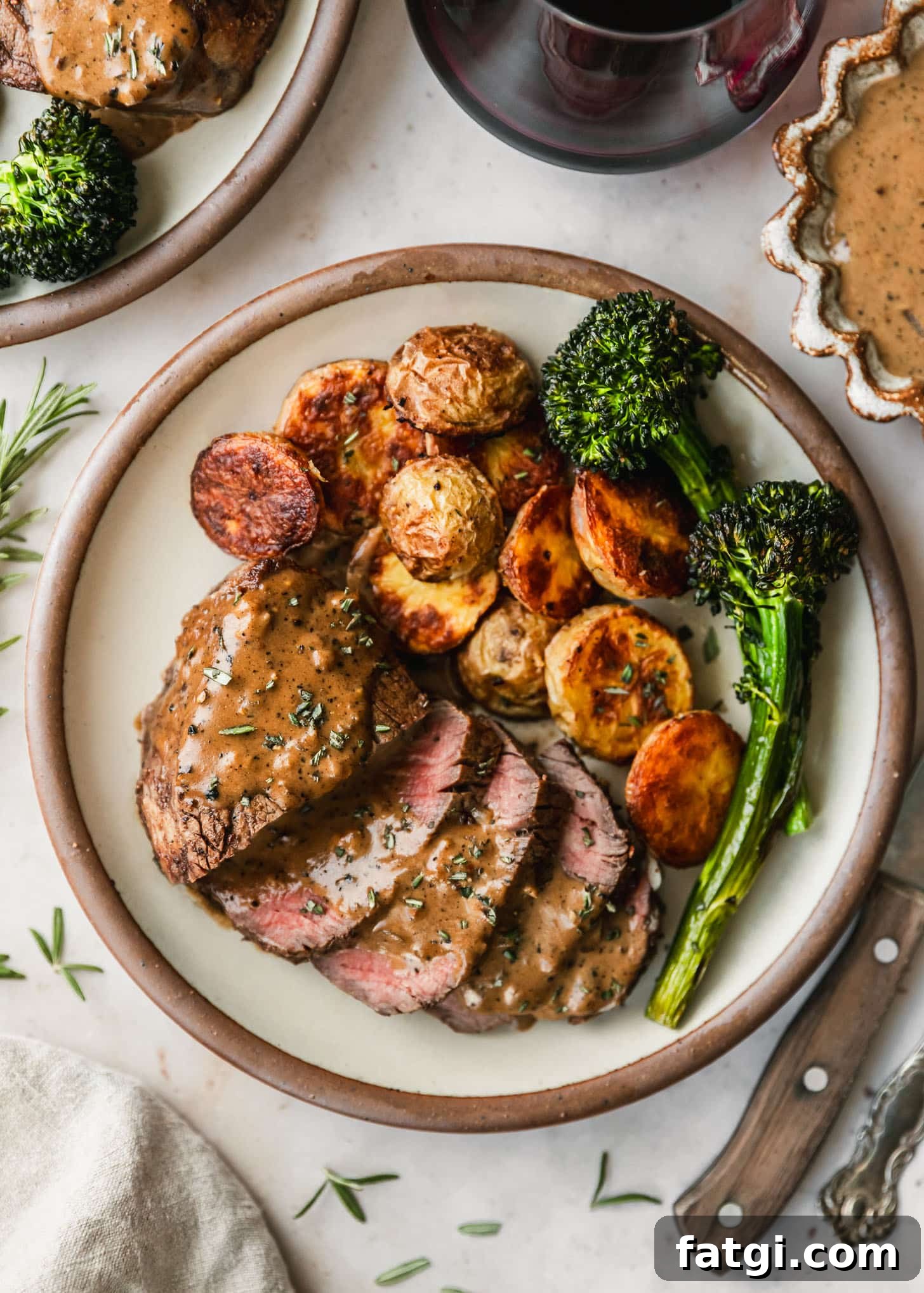 Reverse-Seared Filet Mignon with Creamy Peppercorn Sauce 8 Reverse sear steak, potatoes, and broccolini on a white plate next to red wine, a brown bowl of peppercorn sauce, and a wood steak knife on a marble counter.