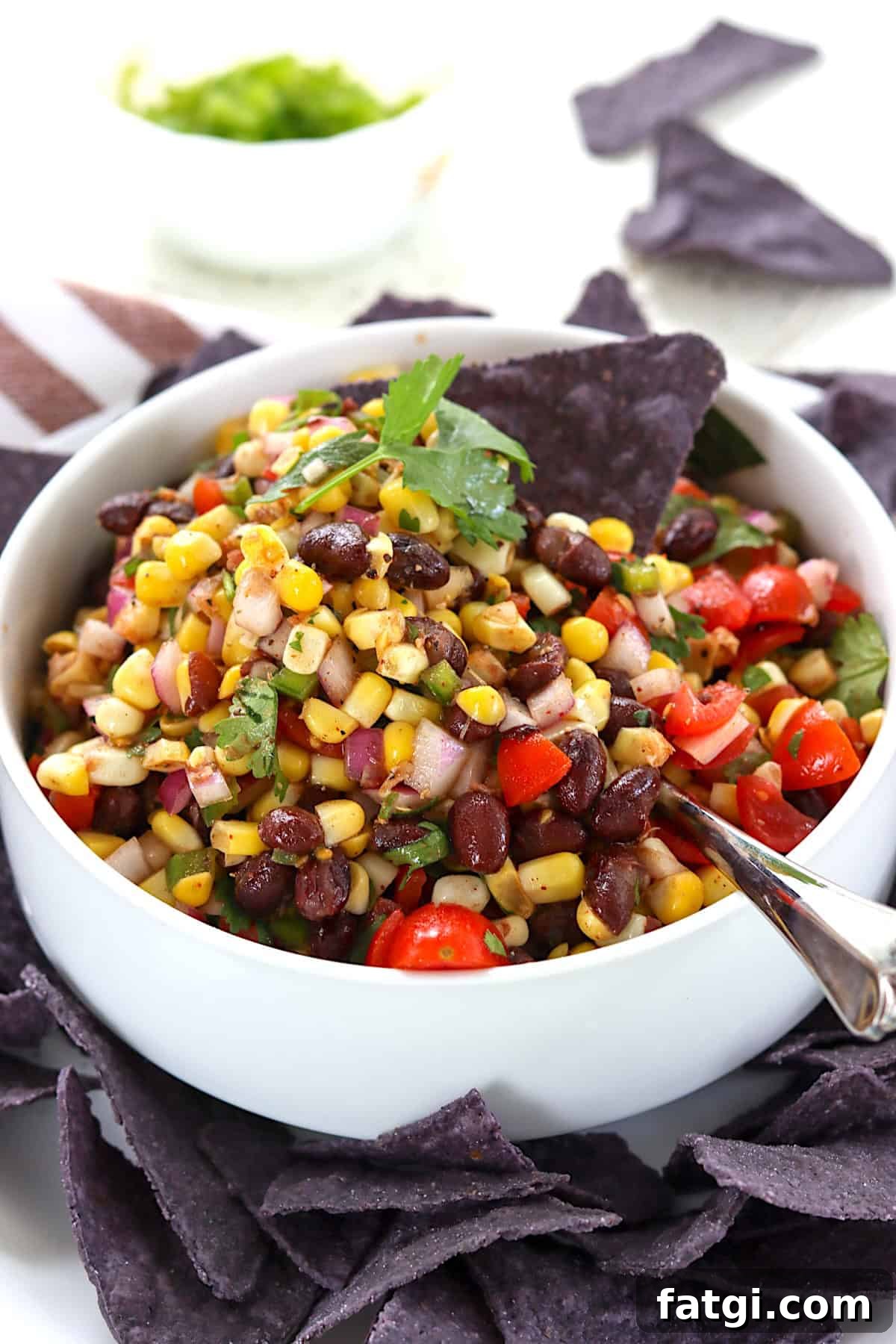 A white serving bowl with black bean and corn salsa with a spoon and tortilla chips.