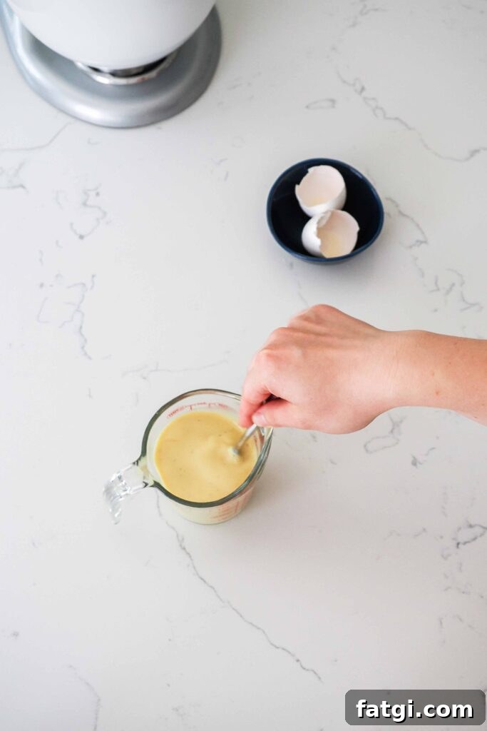 A hand whisks eggs, milk, and vanilla bean paste together in a measuring cup.