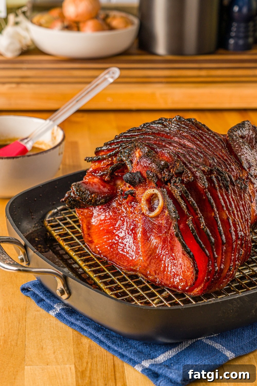 Spiral Ham with Pineapple and Brown Sugar Glaze 2 A baked glazed spiral ham in a roasting pan lined with a rack. The pan is on a wooden counter with a blue cloth napkin and small bowl of glaze with a basting brush.