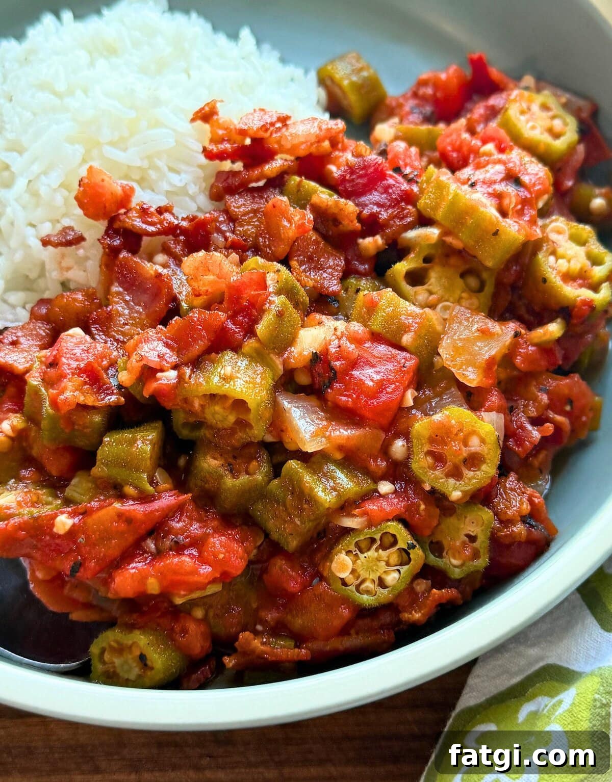 A plate featuring hearty stewed okra and tomatoes with bacon paired with a generous portion of fluffy white rice, highlighting vibrant red tomatoes and green okra pieces.