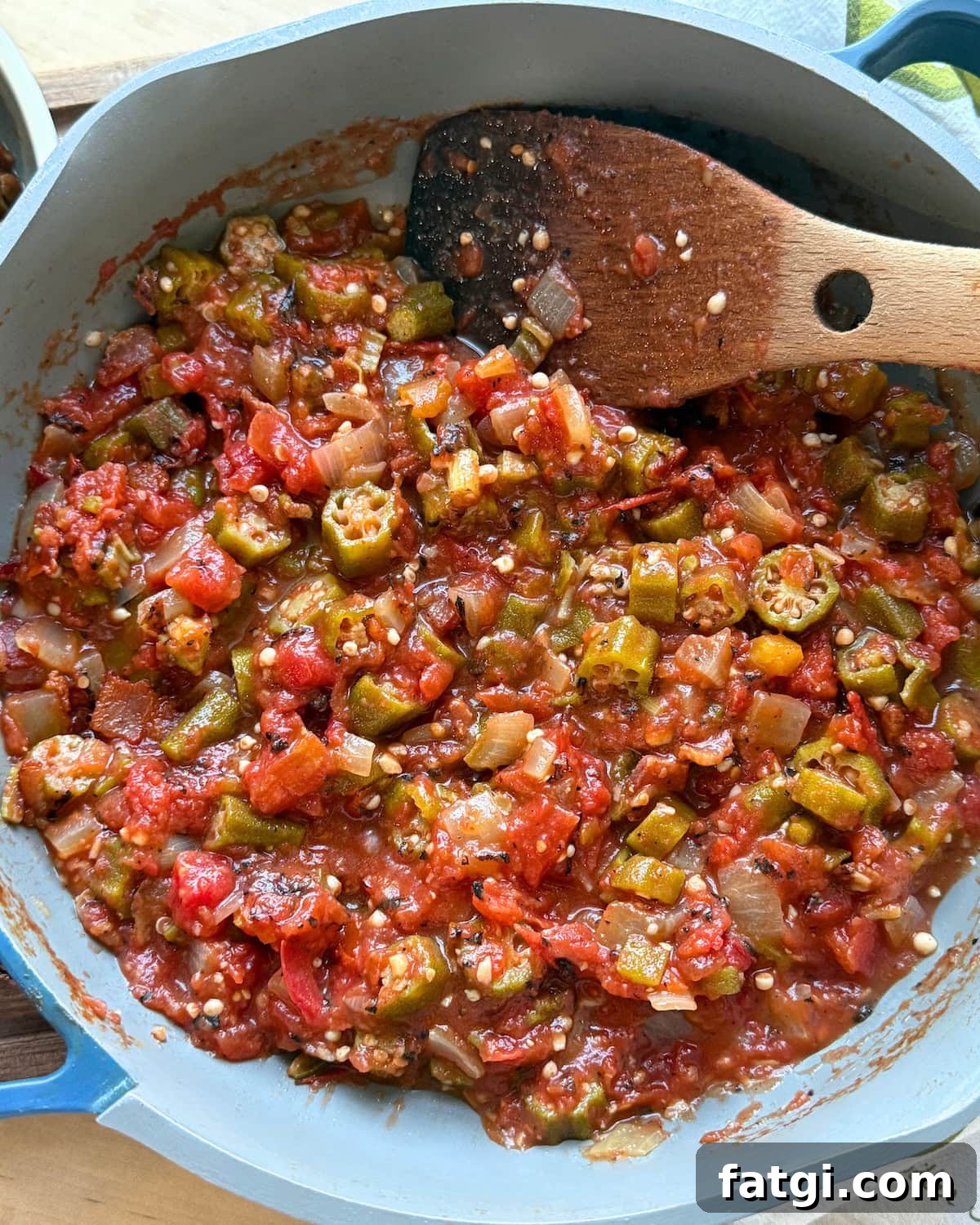 A close-up of a pot containing a colorful mixture of diced okra, tomatoes, onions, and spices, with a wooden spoon resting on the side.