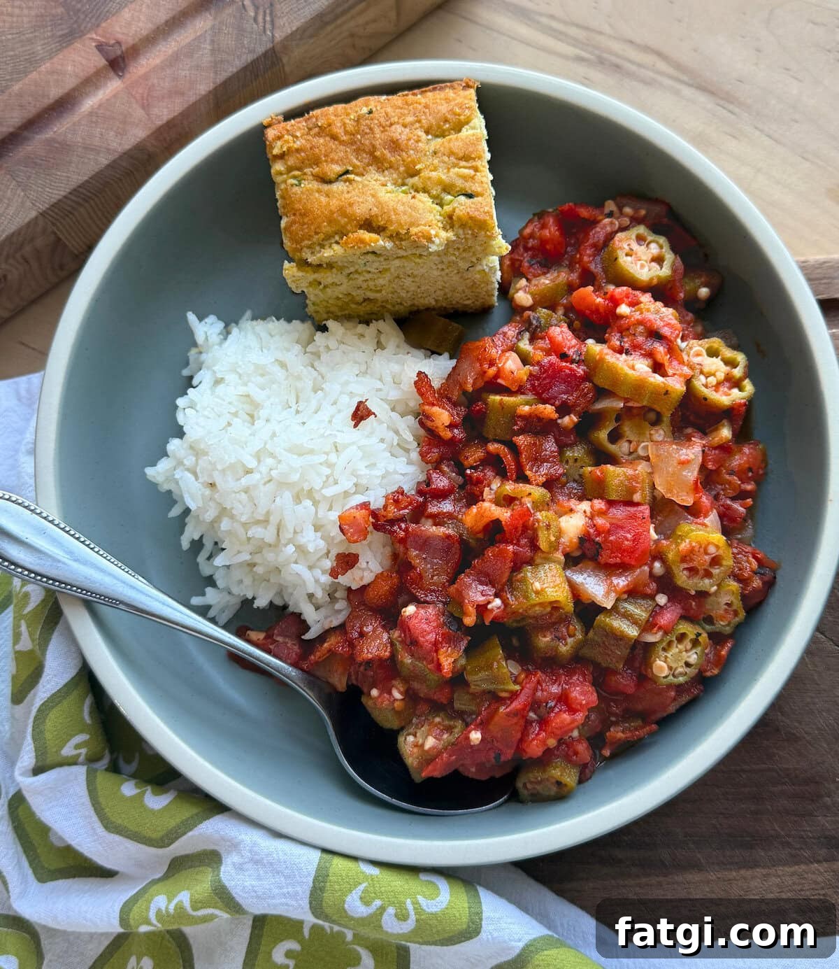 A serving of okra stew with tomatoes and a side of fluffy rice, accompanied by a piece of cornbread in a bowl, set on a wooden surface.