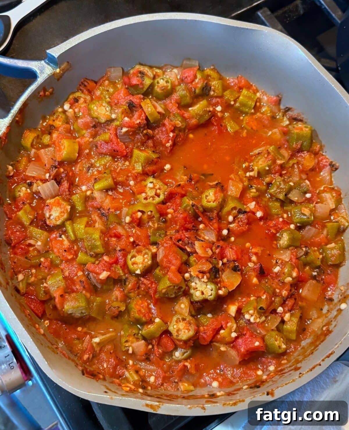 A close-up view of a cooked vegetable medley featuring okra, diced tomatoes, and onions in a vibrant red sauce, simmering in a large pot on the stovetop.
