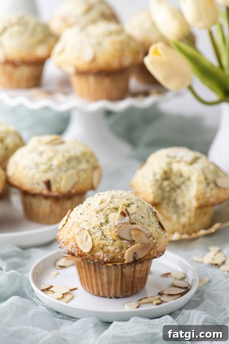 Almond Poppy Seed Muffins Recipe 2 A poppy seed muffin topped with sliced almonds sits on a small white plate. More muffins and a bouquet of yellow tulips are blurred in the background.
