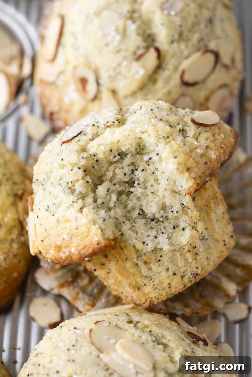 Almond Poppy Seed Muffins Recipe 3 A close-up of poppy seed muffins topped with sliced almonds. One muffin is broken in half, showing its soft, moist texture. The muffins rest on a metal tray with scattered almond slices.
