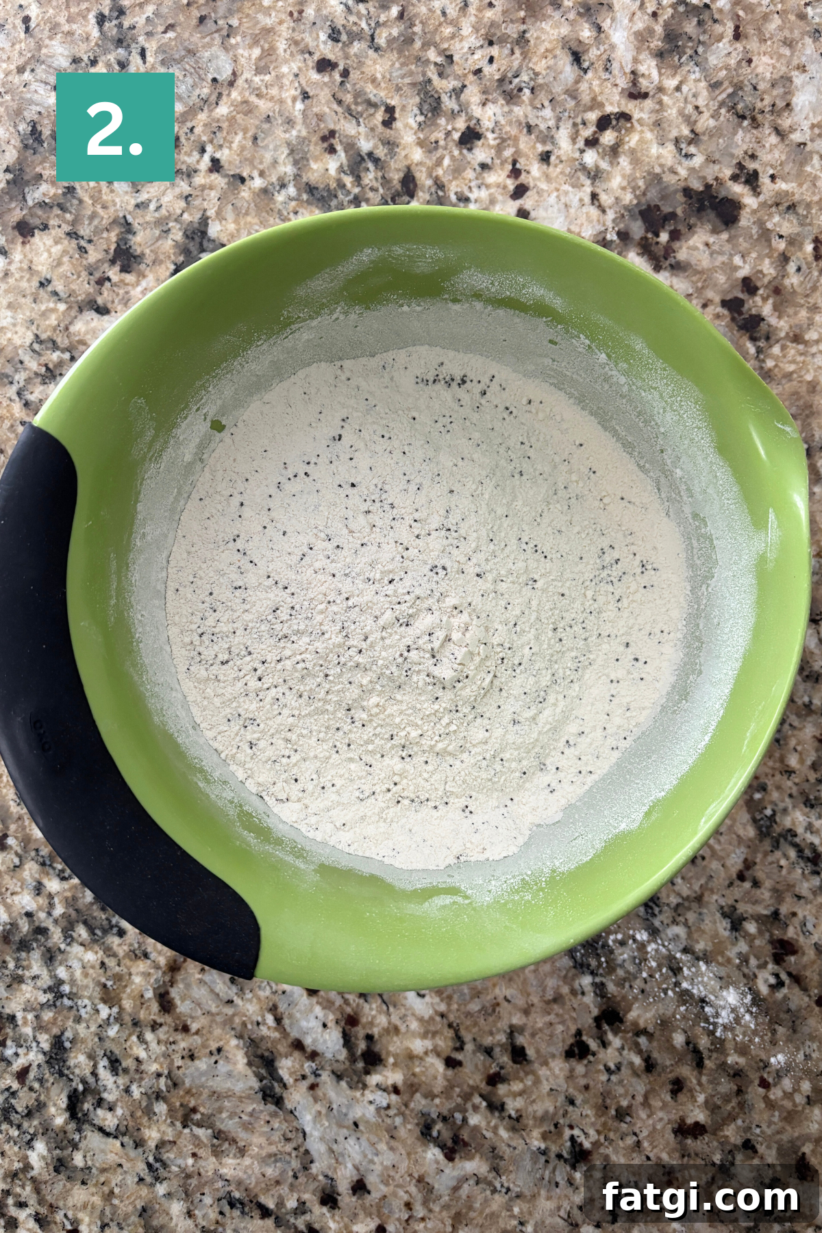 Almond Poppy Seed Muffins Recipe 6 A green mixing bowl with black grip holds a mixture of flour and specks of seasoning on a granite countertop.