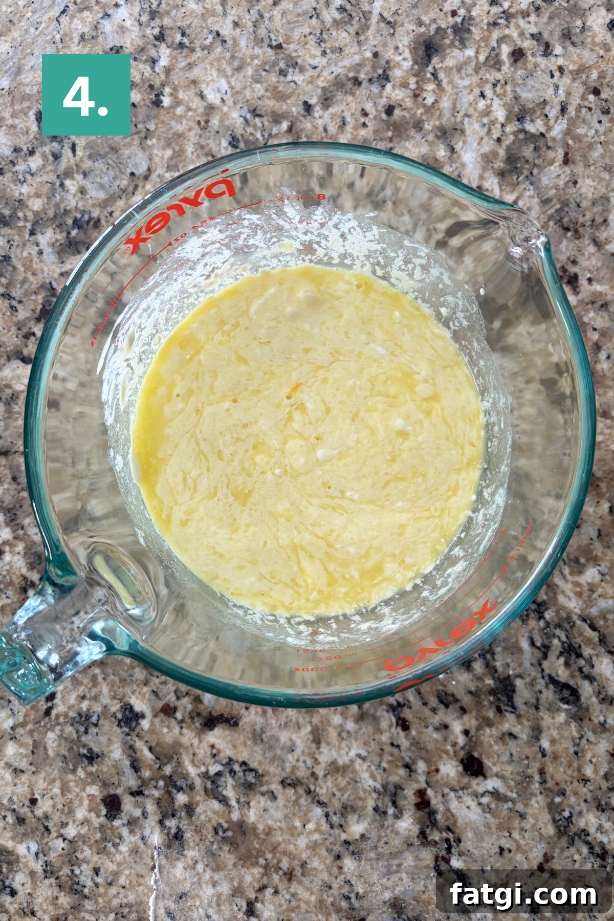 Almond Poppy Seed Muffins Recipe 8 A glass measuring cup with a yellow, slightly lumpy batter mixture sits on a granite countertop.