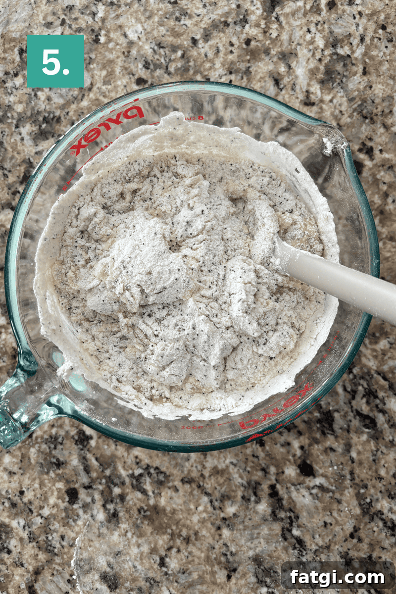Almond Poppy Seed Muffins Recipe 9 A glass measuring cup filled with flour mixture and a white spatula sits on a granite countertop.