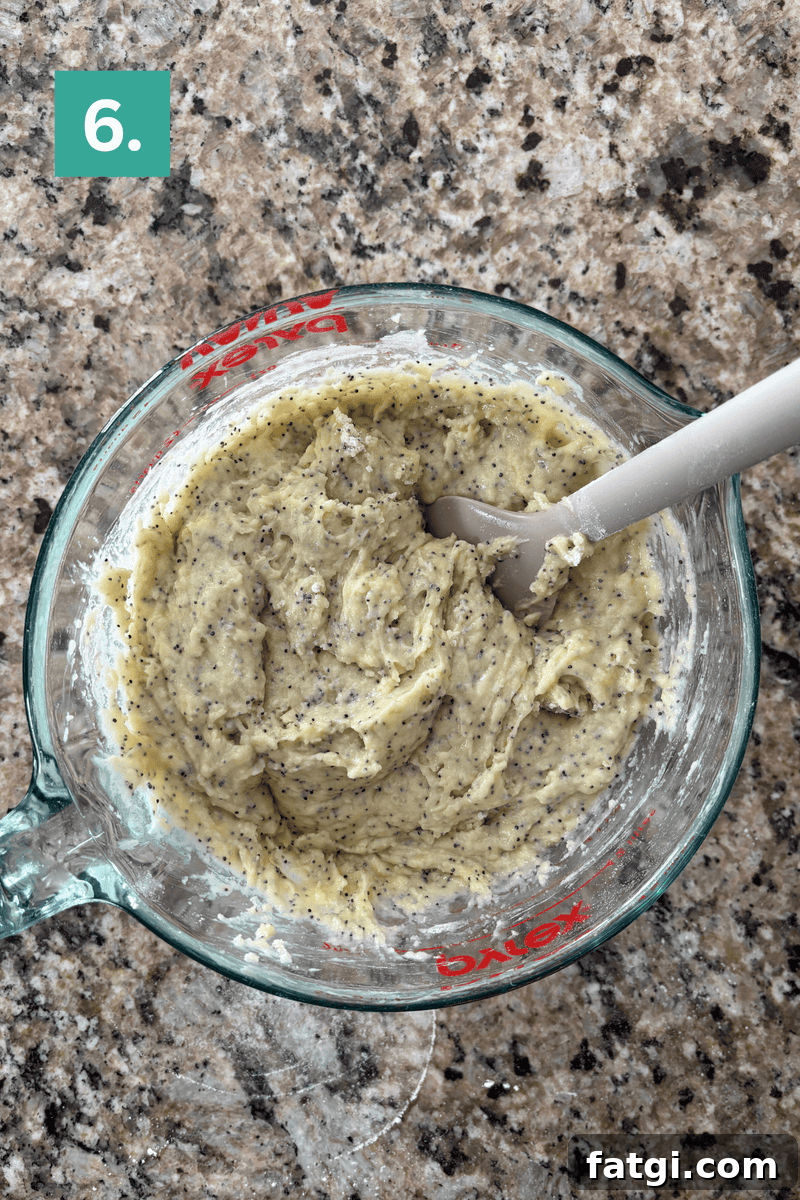 Almond Poppy Seed Muffins Recipe 10 A glass measuring cup filled with a thick, pale batter mixed with poppy seeds is being stirred with a gray spatula.