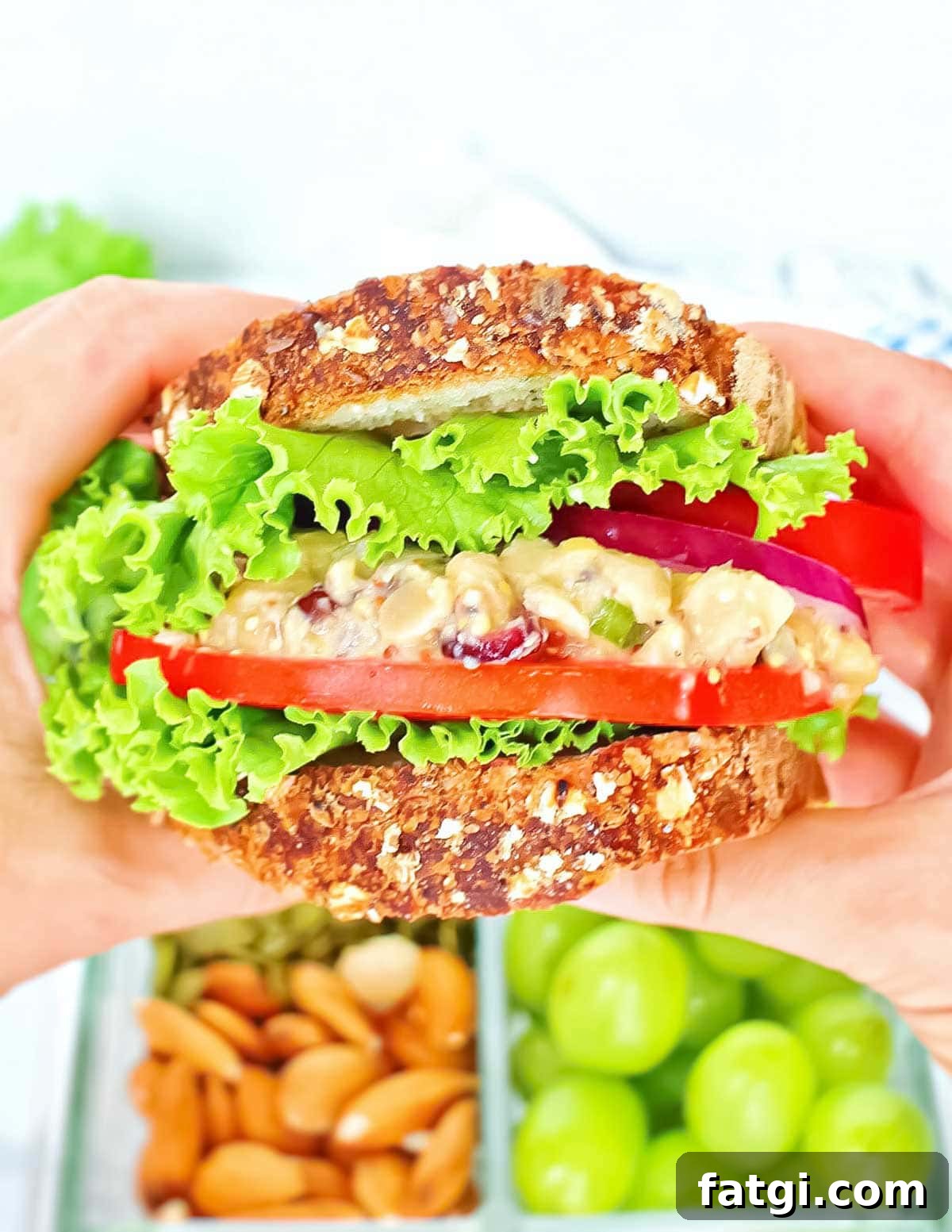 Two hands holding a sandwich with lettuce, tomatoes, onion, and chickpea salad.