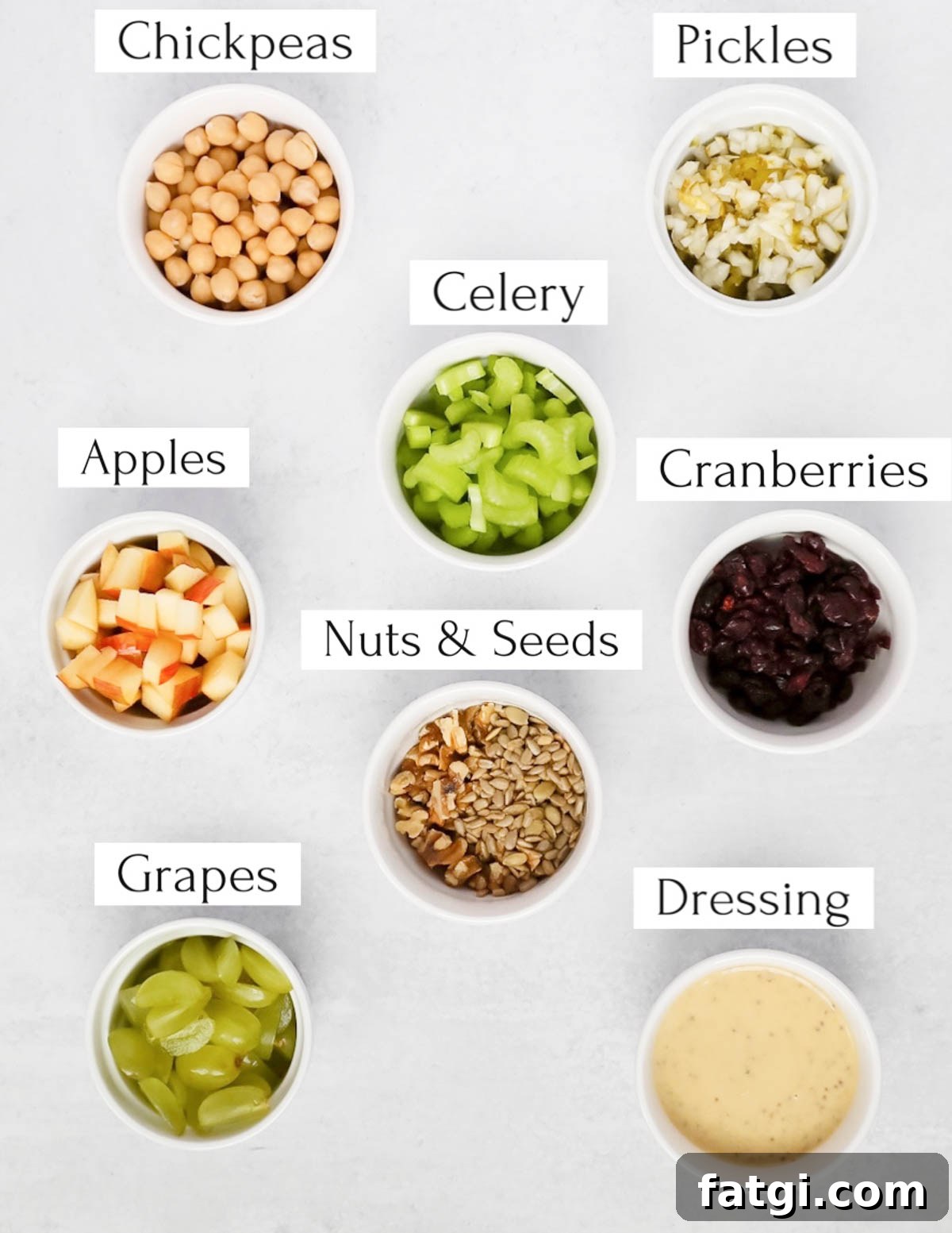 Labeled ingredients in small white bowls including chickpeas, pickles, celery, apples, cranberries, nuts and seeds, grapes, and dressing.