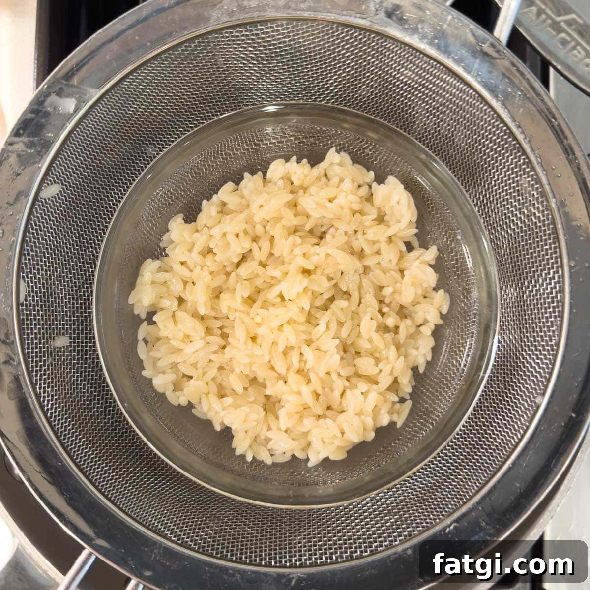 Cooked orzo pasta in a sieve over a bowl being rinsed and drained.