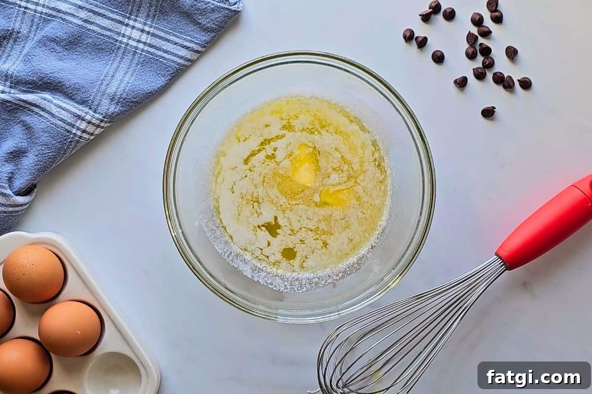 Stick of butter nearly fully melted in glass bowl next to whisk.