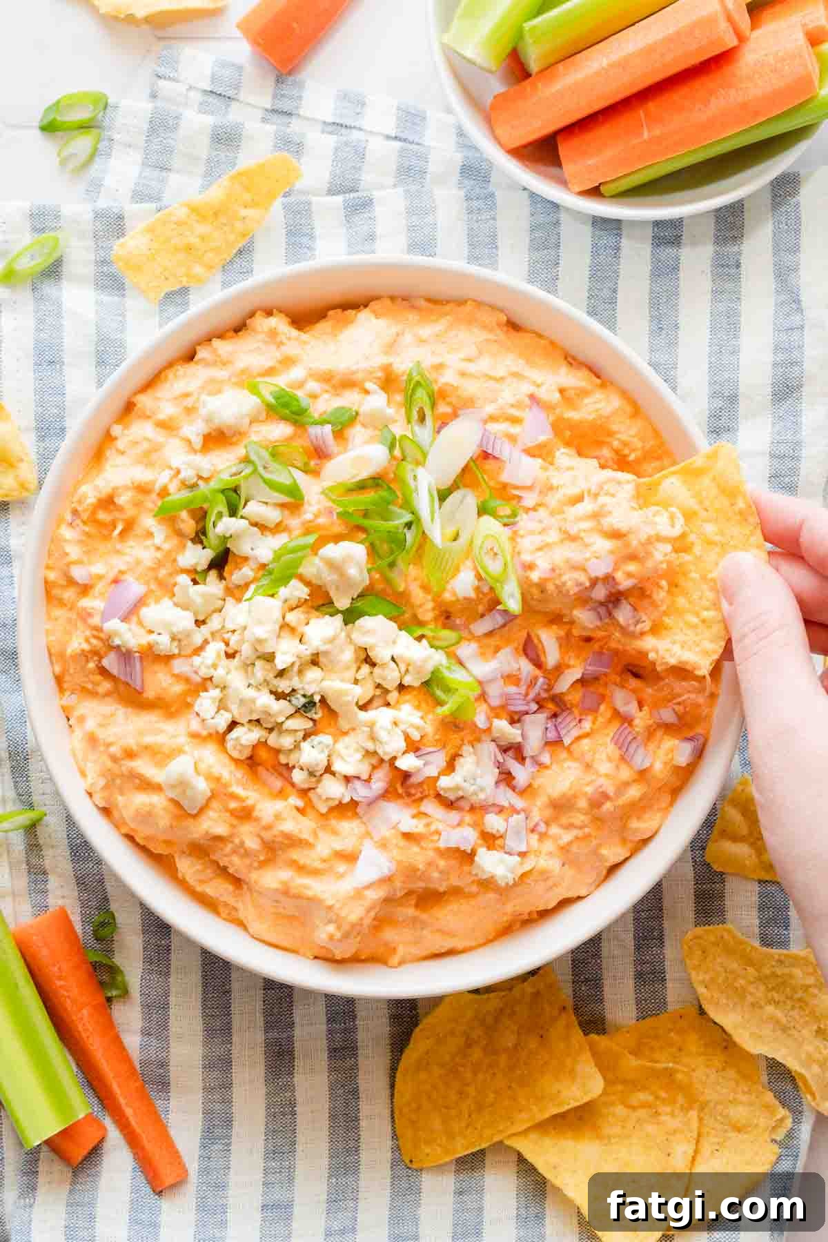 hand scooping chip into bowl of buffalo chicken dip with veggies and chips on the side.